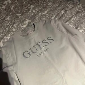Grå långärmad tröja från Guess - Snygg grå långärmad tröja från Guess med tryck 'EST. 1981' på framsidan. Tröjan har en slim fit passform och är perfekt för en stilren look. Tillverkad i mjukt material för extra komfort. Storlek L men passar m/s
