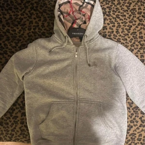 Grå hoodie från Tenson - Säljer en grå hoodie från Tenson med dragkedja och en klassisk rutmönstrad insida i huvan. Perfekt för en avslappnad stil. Den har långa ärmar och en praktisk ficka framtill.