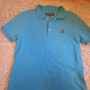 Turkos pikétröja från Lyle & Scott - Snygg turkos pikétröja från Lyle & Scott i 100% bomull. Tröjan har en klassisk krage med knappar och det ikoniska broderade logotypmärket på bröstet. Tröjan är i en unik färg.