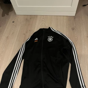 Svart träningströja  från Adidas - Snygg svart träningströja från Adidas med vita ränder längs ärmarna och Tysklands landslagsemblem på bröstet. Jackan har dragkedja och långa ärmar, perfekt för sportiga aktiviteter. Dragkedjan lite sönder kolla bild 3!