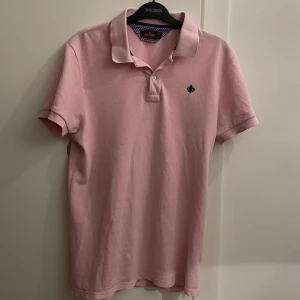 pikétröja från Morris - Snygg rosa pikétröja från Morris med en liten broderad detalj på bröstet. Tröjan har en klassisk krage och knappar framtill. Perfekt för en stilren och avslappnad look.