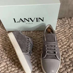 Grå sneakers från Lanvin - Snygga grå sneakers från Lanvin med en kombination av mocka och glansigt material. Skorna har en klassisk snörning och en vit sula. Perfekta för en stilren look.