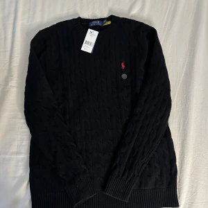 Svart stickad tröja från Polo Ralph Lauren - Snygg svart stickad tröja från Polo Ralph Lauren med det klassiska röda logotypbroderiet på bröstet. Tröjan är tillverkad i 100% bomull och har en ribbad rund halsringning samt långa ärmar. Perfekt för en stilren look.