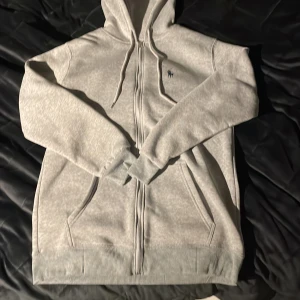 Grå hoodie från Ralph Lauren - Säljer en stilren grå hoodie från Ralph Lauren med dragkedja och broderad logga på bröstet. Tröjan har långa ärmar och en praktisk huva med snörning. Perfekt för en avslappnad look.