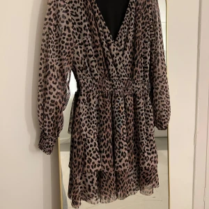 Leopardmönstrad klänning med v-ringning - Trendig leopardmönstrad klänning med v-ringning och långa ärmar. Klänningen har en smickrande omlott-design och elastisk midja för en bekväm passform. 