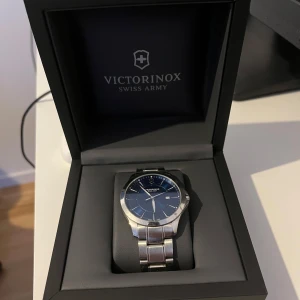 Silverfärgad klocka från Victorinox - Elegant klocka från Victorinox med en stilren blå urtavla och datumvisning. Armbandet är i silverfärgat stål och klockan levereras i originalförpackning. Perfekt för den som söker en tidlös accessoar. Pris kan diskuteras. Man kan såklart ändra storlek. Köpte den för 6000kr 