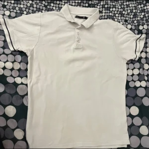 Vit kortärmad pikétröja - Säljer en klassisk vit pikétröja med korta ärmar och knappar framtill. Perfekt för en stilren och avslappnad look. Tröjan har en krage och diskreta ränder på ärmsluten.