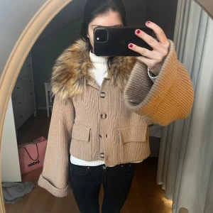 Beige stickad jacka med fuskpäls - Snygg beige stickad jacka med stor krage i fuskpäls. Jackan har knappar framtill och två fickor. Perfekt för kyliga dagar när du vill vara både varm och stilren.