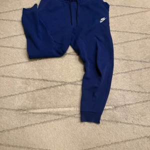 Blå mjukisbyxor från Nike - Snygga blå mjukisbyxor från Nike med elastisk midja och dragsko. Byxorna har en avslappnad passform och är perfekta för en chill dag. Nike-loggan i vitt på sidan ger en sportig touch.