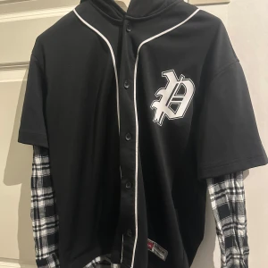 Svart kortärmad tröja med rutiga ärmar - Snygg svart kortärmad tröja som har luva,  med vita detaljer och ett stort tryck på bröstet. Tröjan har knappar framtill och är kombinerad med rutiga långärmade ärmar i svart och vitt. Perfekt för en unik och stilren look.