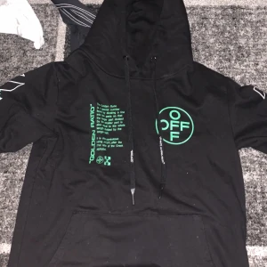 Svart hoodie från Off-White - Snygg svart hoodie från Off-White med gröna detaljer och tryck både fram och bak. Den har en justerbar huva med snören och en stor ficka framtill. Perfekt för en streetwear-look.