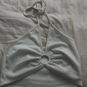 Vit topp med en fin detalj  - Snygg vit överdel med knytband i nacken och en dekorativ ringdetalj framtill. Perfekt för en dag på stranden eller vid poolen.