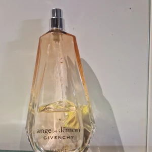 Ange ou Démon Le Secret från Givenchy 100 ml - Elegant parfymflaska med en unik, droppformad design. Flaskan är genomskinlig med en ljusrosa nyans och har en silverfärgad spraytopp. Texten 'ange ou démon' och 'le secret' är tryckt på framsidan. Perfekt för den som söker en sofistikerad doftupplevelse.  Kvar av parfym: ca 50 procent 