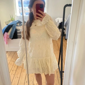 Gul blommig klänning med öppen rygg - Charmig gul klänning med blommigt mönster och volangdetaljer. Den har långa ärmar och en elegant öppen rygg med knytning. Perfekt för vår och sommar.