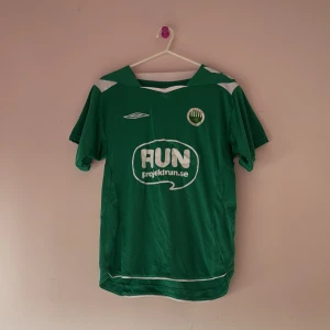 Grön fotbollströja från Umbro - Säljer en grön fotbollströja från Umbro med vit logga och sponsortryck på framsidan. Tröjan har korta ärmar och en klassisk krage. Perfekt för fotbollsfans eller träning!