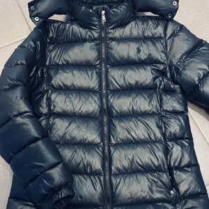 Svart tunnare dunjacka från Polo Ralph Lauren - Snygg svart dunjacka från Polo Ralph Lauren med broderad logotyp på bröstet. Jackan har en hög krage och dragkedja framtill, perfekt för kyliga dagar. Den quiltade designen ger både stil och värme. Storlek XL (junior), 16 år. Figursydd modell. 