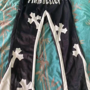 Svarta flared byxor med vita korsmönster - Unika svarta byxor med vita korsmönster och textdetaljer. Byxorna har en loose passform och är perfekta för en avslappnad stil. De är tillverkade i ett syntetmaterial som ger en skön känsla.