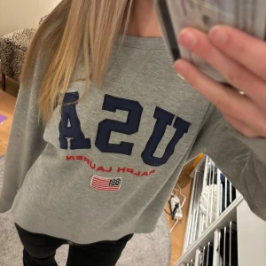 Grå Ralph Lauren tröja - Säljer en grå sweatshirt från Ralph Lauren med texten 'USA' i blått och en liten amerikansk flagga. Tröjan har långa ärmar och en rund halsringning. Tröjan är i storlek Xs och säljer pga att den ej kommer till användning.