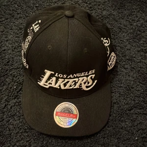 Svart keps från Mitchell & Ness - Snygg svart keps med Los Angeles Lakers-broderi framtill. Kepsen har justerbar passform och flera broderade detaljer runtom. Perfekt för sportfantasten!