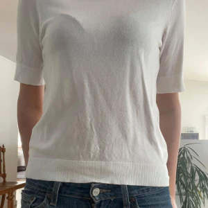 Vit kortärmad tröja - Säljer en stilren vit tröja med korta ärmar. Perfekt för en avslappnad look. Tröjan har en ribbad kant vid ärmslut och nederkant.