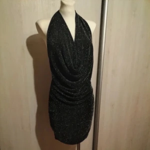 Svart glittrig klänning - Snygg svart glittrig klänning med draperad framsida och halterneck-design. Perfekt för en kväll ute med stil. Klänningen har en åtsittande passform som framhäver figuren. Skicka om du har frågor!
