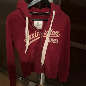 Röd hoodie från Lexington - Säljer en snygg röd hoodie från Lexington med dragkedja och vit text 'Lexington 1993' på framsidan. Den har en justerbar huva med vita snören och praktiska fickor framtill.