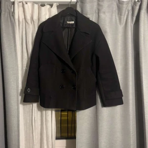 Svart kappa från Design by Si - Elegant svart kappa med dubbelknäppning och breda slag. Jackan har långa ärmar och en klassisk design som passar perfekt för kyligare dagar. Tillverkad i ett stilrent material som ger en sofistikerad look. Kappan är i storlek XS/S!! Pris kan diskuteras