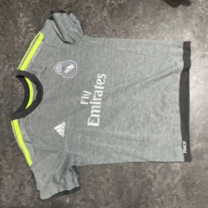 Grå fotbollströja från Adidas - Snygg grå fotbollströja från Adidas med korta ärmar och detaljer i svart och neongul. Tröjan har en rund halsringning och klubbmärke på bröstet. Perfekt för fotbollsfans!