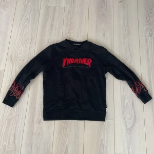 Svart tröja från Vans med Thrasher-tryck - Säljer en svart tröja från Vans med Thrasher-tryck i rött på framsidan. Tröjan har långa ärmar med röda flammor som detaljer. Perfekt för en cool och avslappnad stil. 'Off the Wall' text finns på insidan av kragen. 