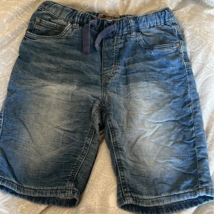 Blå jeansshorts från Lab Industries för barn  - Snygga blå jeansshorts från Lab Industries med elastisk midja och snörning. De har en avslappnad stil med fickor fram och bak, perfekt för en casual look. Märkeslogga broderad på bakfickan.