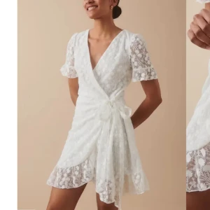 Vit spetsklänning med volangdetaljer - Elegant vit klänning i spets med korta ärmar och omlott-design. Klänningen har en v-ringad hals och knytband i midjan för en smickrande passform. Volangdetaljer vid ärmslut och nederkant ger en romantisk touch.