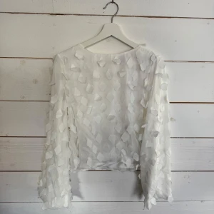 Vit blus med 3D-detaljer - Säljer en elegant vit blus med unika 3D-detaljer som ger en lyxig känsla. Blusen har långa ärmar och en knapp i nacken för enkel påklädning. Finns matchande kjol till.