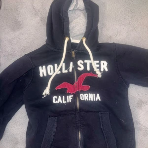 Svart hoodie från Hollister - Säljer en svart hoodie från Hollister med vit text och en röd logotyp på framsidan. Den har en dragkedja och justerbara snören i huvan. Storlek S och defekter förekommer!❤️