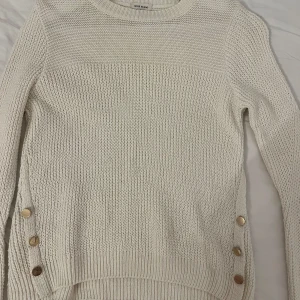 Vit stickad tröja från River Island - Säljer en stilren vit stickad tröja från River Island. Tröjan har långa ärmar och dekorativa guldknappar på sidorna. Perfekt för kyligare dagar och ger en elegant touch till din outfit.