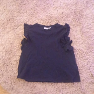 Mörkblå topp från Gina Tricot - Säljer en mörkblå topp från Gina Tricot med volangdetaljer vid ärmarna. Toppen är ärmlös och har en enkel design som passar perfekt till vardags. Perfekt för dig som gillar en stilren look.