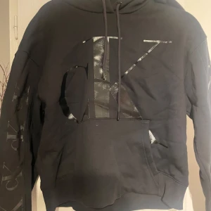 Svart hoodie från Calvin Klein Jeans - Säljer en stilren svart hoodie från Calvin Klein Jeans med stor logga på framsidan. Hoodien har en klassisk känguruficka och justerbar huva med dragsko. Perfekt för en avslappnad look.