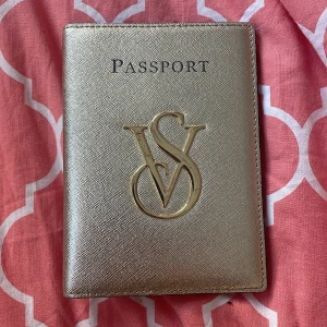 Guld passfodral från Victoria's Secret - Snyggt passfodral i guld från Victoria's Secret med en elegant logotyp på framsidan. Insidan är klädd i en härlig rosa färg och har fack för både pass och boardingkort. Perfekt för att resa med stil!