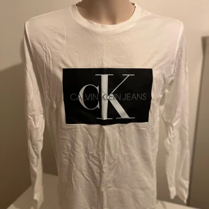 Vit långärmad tröja från Calvin Klein - Säljer en stilren vit långärmad tröja från Calvin Klein Jeans med ett stort svart tryck på framsidan. Perfekt för en avslappnad och trendig look. Tröjan är gjord i mjukt bomullsmaterial och har en klassisk passform.