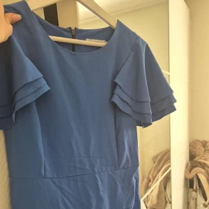 Blå klänning med volangärmar - Säljer en elegant blå klänning med volangdetaljer på ärmarna. Klänningen har en figurnära passform och en dragkedja i ryggen. Perfekt för speciella tillfällen!