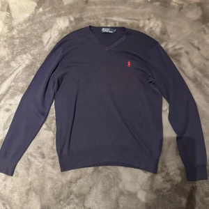Mörkblå tröja från Polo Ralph Lauren - Säljer en stilren mörkblå tröja från Polo Ralph Lauren med v-ringning och det klassiska röda logotypbroderiet på bröstet. Tröjan har långa ärmar och är tillverkad i ett mjukt material som passar perfekt för kyligare dagar.