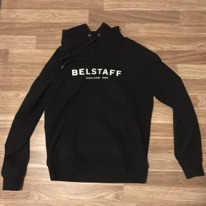 Svart hoodie från Belstaff - Säljer en stilren svart hoodie från Belstaff med vit logotyp på bröstet. Hoodien har en justerbar dragsko i huvan och en praktisk magficka. Perfekt för en avslappnad look. Nypris ca 999kr