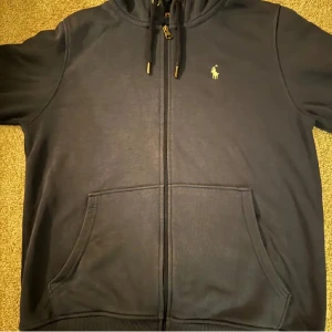 Svart hoodie från Ralph Lauren - Säljer en stilren svart hoodie från Ralph Lauren med dragkedja och det klassiska logotypbroderiet på bröstet. Tröjan har en justerbar huva med snörning och praktiska fickor framtill. Perfekt för en avslappnad look.