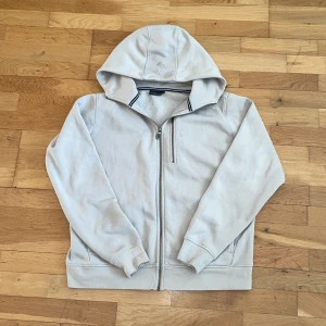 Beige huvtröja från Sail Racing - Säljer en beige huvtröja från Sail Racing med dragkedja och en bröstficka. Tröjan har en mjuk insida och är tillverkad i en bomulls- och polyesterblandning. Perfekt för kyliga dagar med sin bekväma passform och stilrena design.