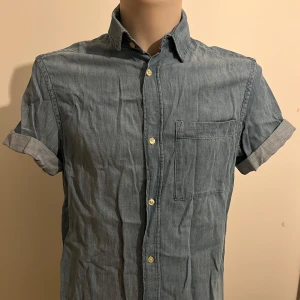 Blå jeansskjorta från H&M - Snygg blå jeansskjorta från H&M med korta ärmar och klassisk krage. Skjortan har knappar framtill och en bröstficka för en avslappnad look. Perfekt för en casual stil.