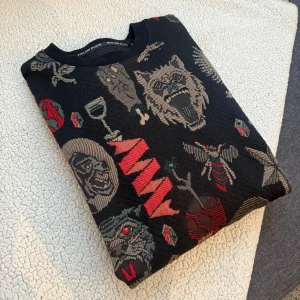 Svart mönstrad tröja från Philipp Plein - Säljer en svart tröja från Philipp Plein med ett unikt mönster av vargar, dödskallar och andra grafiska element i beige och rött. Tröjan är tillverkad i 100% bomull och har långa ärmar med ribbade muddar.
