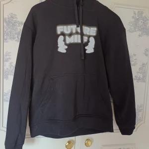 Svart hoodie med tryck - Säljer en svart hoodie med texten 'FUTURE MILF' tryckt på framsidan. Hoodien har en klassisk design med känguruficka och justerbar huva med dragsko. Perfekt för en avslappnad stil.