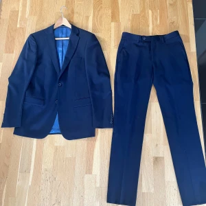Mörkblå kostym (Dressmann) - Elegant mörkblå kostym med kavaj och byxor. Kavajen har två knappar och en klassisk passform. Byxorna är stilrena med en knapp och dragkedja. Perfekt för formella tillfällen. Kavajen är storlek 46 (lång slim fit) och byxorna är 30/34 (slim fit). 45% ull. Hör av er vid frågor :)