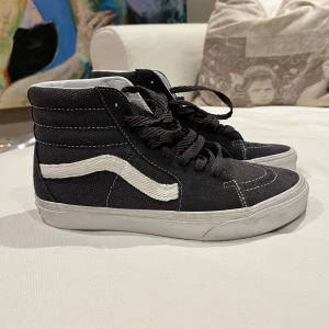 Svarta sneakers från Vans - Säljer ett par klassiska svarta Vans sneakers med vit sula och den ikoniska vita sidostripen. Skorna har snörning och en hög modell som ger extra stöd runt ankeln. Perfekta för en avslappnad stil.