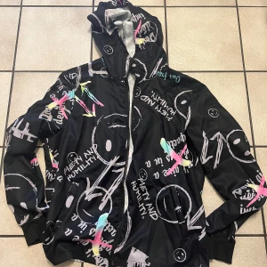 Svart mönstrad jacka med huva storlek M oanvänd  - Cool svart jacka med grafiska mönster i vitt och neonfärger. Jackan har en dragkedja framtill och en praktisk huva. Perfekt för en streetwear-look.