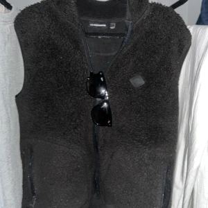 Svart fleeceväst från J.Lindeberg - Säljer en svart fleeceväst från J.Lindeberg. Västen har en dragkedja framtill och är perfekt för kyligare dagar. Den är mjuk och bekväm, vilket gör den till ett bra val för lager-på-lager-stil. Tvättas innan den skickas!!